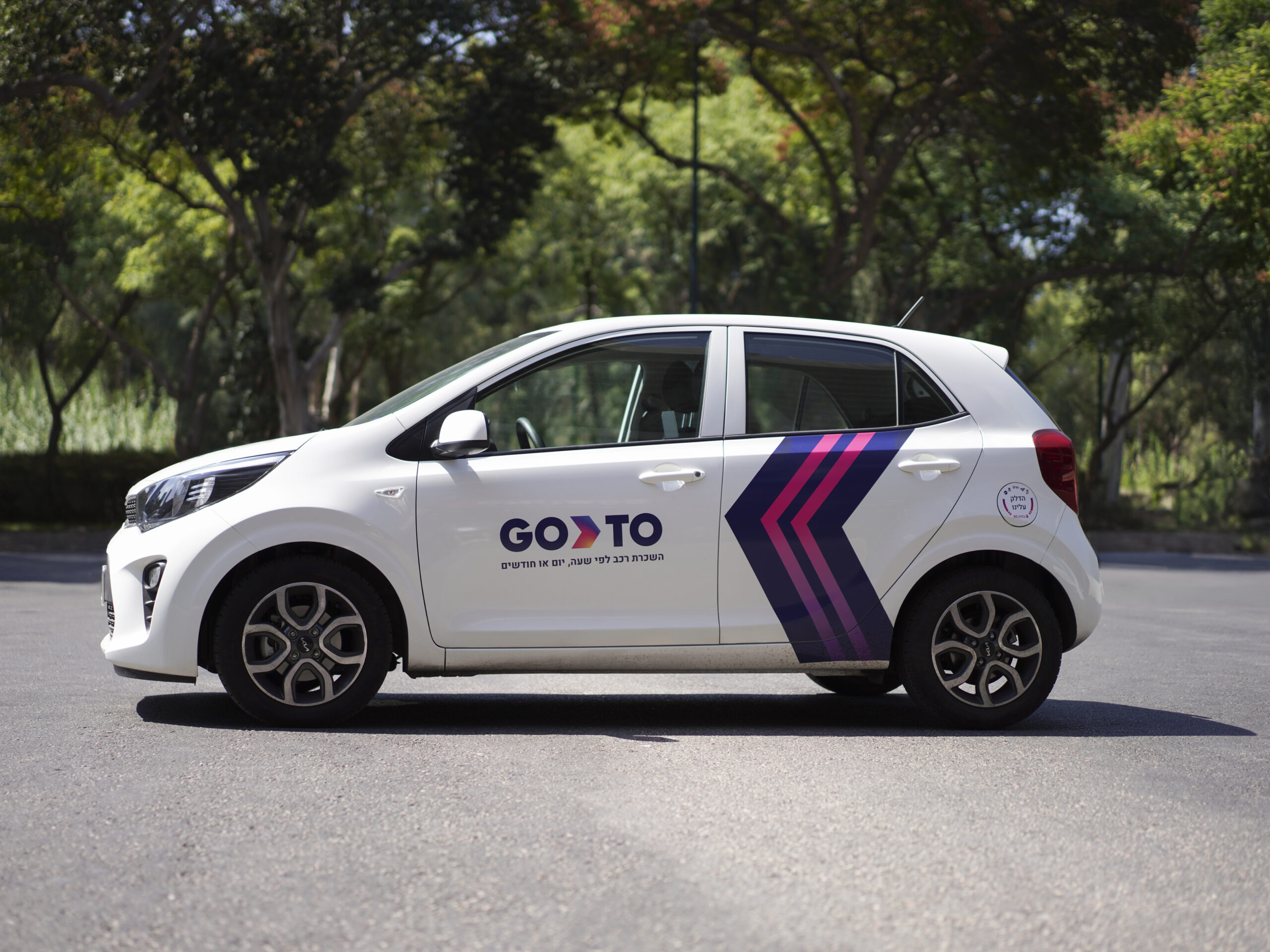 Side view of a white compact car branded with 'GoTo' parked outdoors on a sunny day, promoting the concept of renting a car for a day.