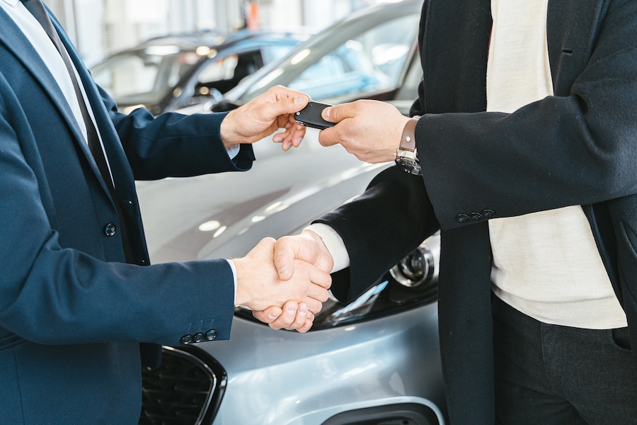 Two business professionals shaking hands and exchanging car keys in front of a vehicle, symbolizing a business vehicle leasing agreement.