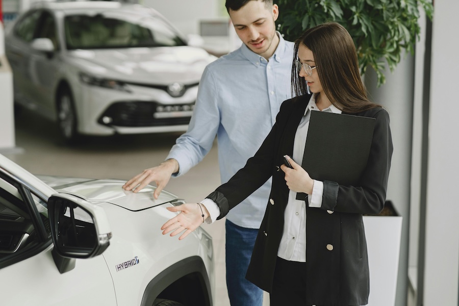A businesswoman and a customer discussing car leasing deal options while reviewing a hybrid vehicle at a dealership.