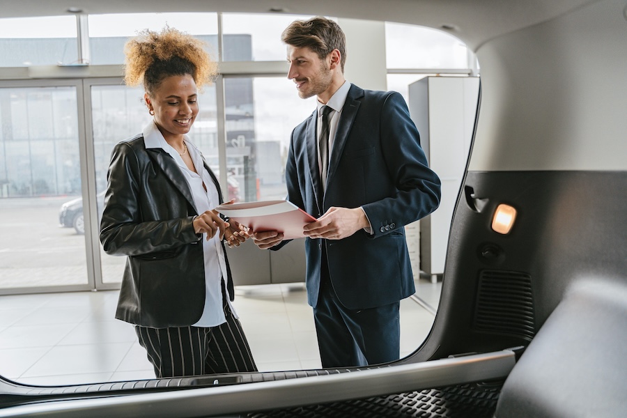 Two business professionals reviewing documents inside a car dealership, discussing company car benefit value and tax implications.